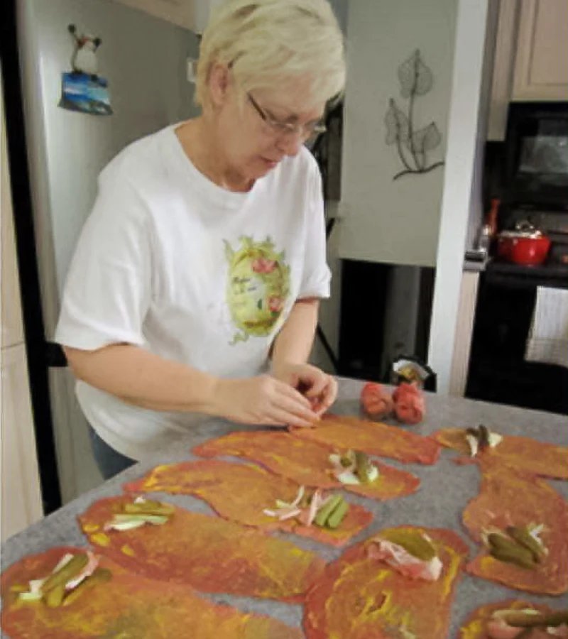Oma assembling German rouladen on a kitchen counter, beef slices topped with bacon, onions, and pickles.