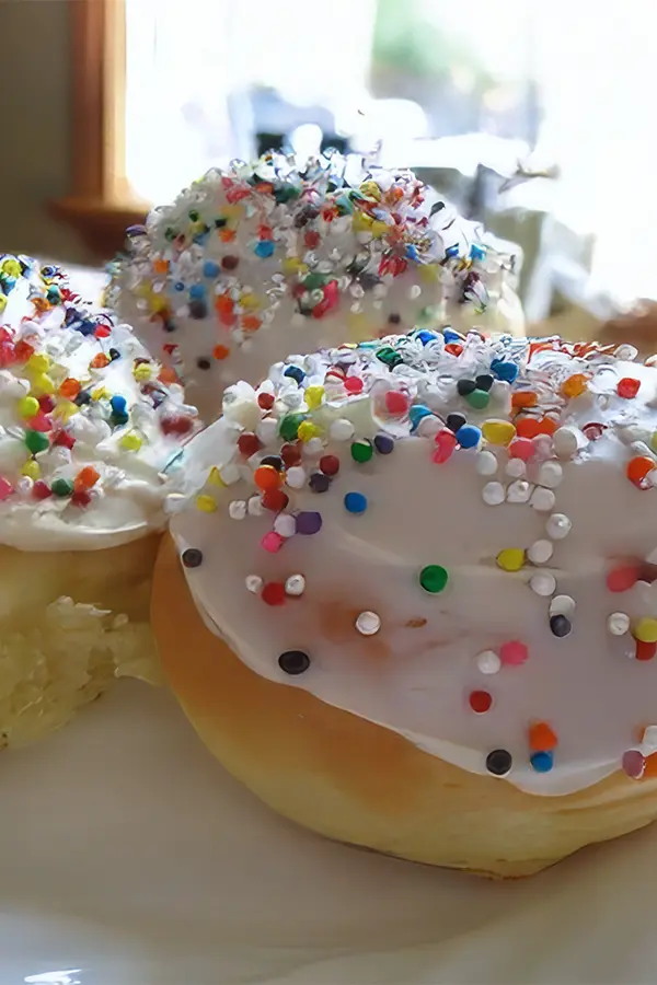 Three frosted paska Easter buns covered in colorful sprinkles are displayed on a plate.