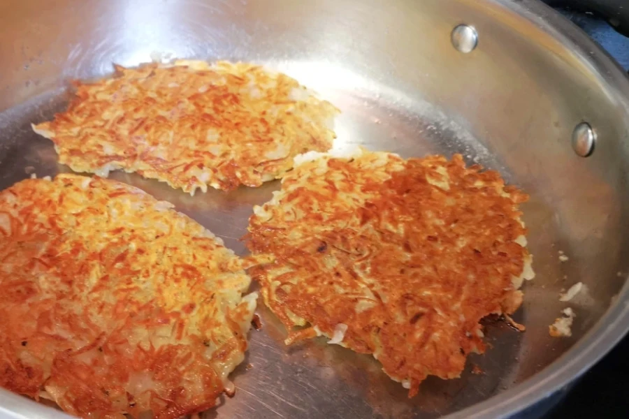Three Kartoffelpuffer (German potato pancakes) frying in a stainless steel pan, an easy German family tradition for a weeknight meal.