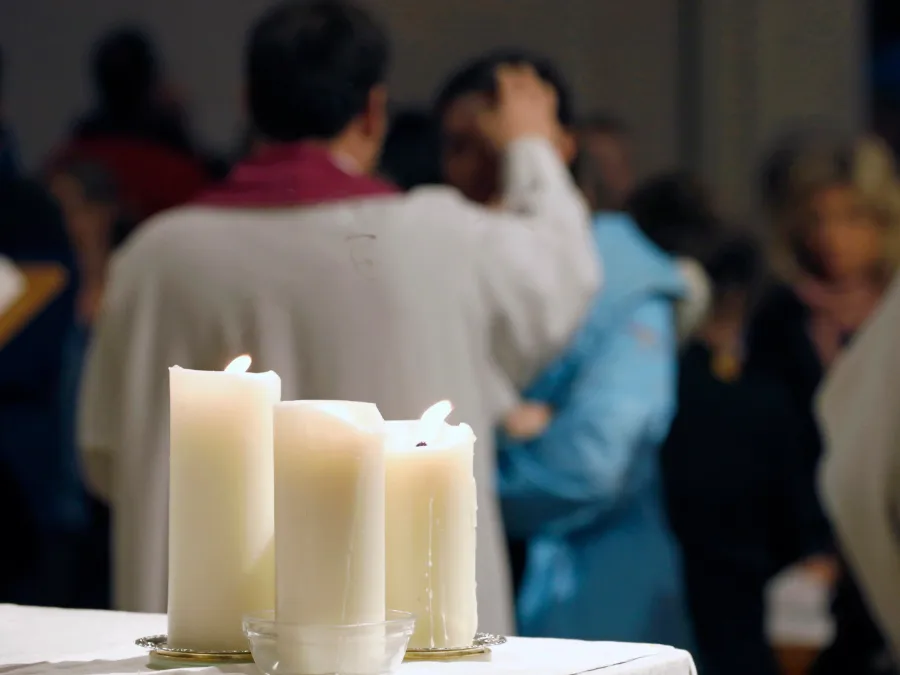 White altar candles in the foreground as a priest marks ashes on a worshipper, Ash Wednesday in Germany.