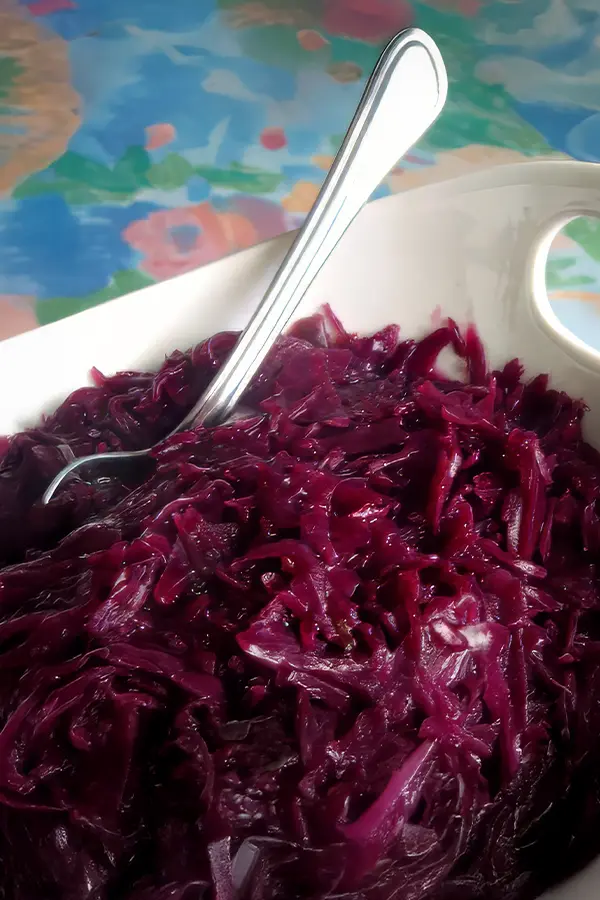 Serving of cooked red cabbage in a white dish with a silver spoon, set against a colorful background.