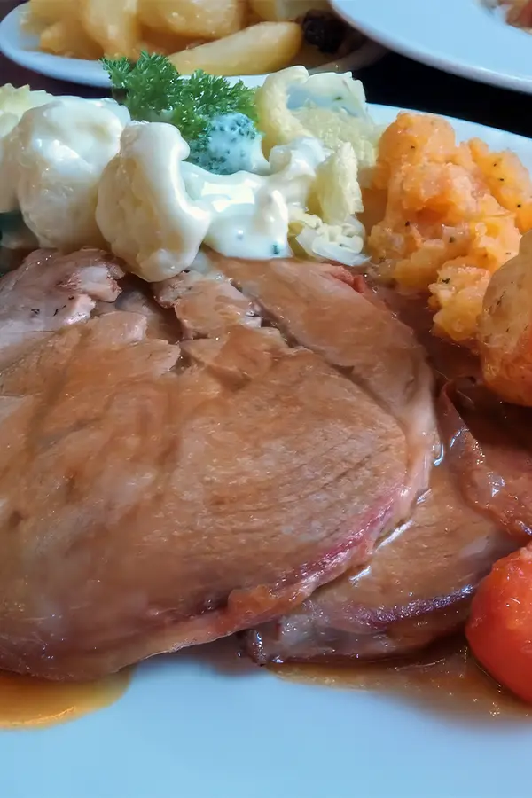 A plate with slices of roast lamb, roasted potatoes, carrots, and a side of creamy cauliflower and broccoli.