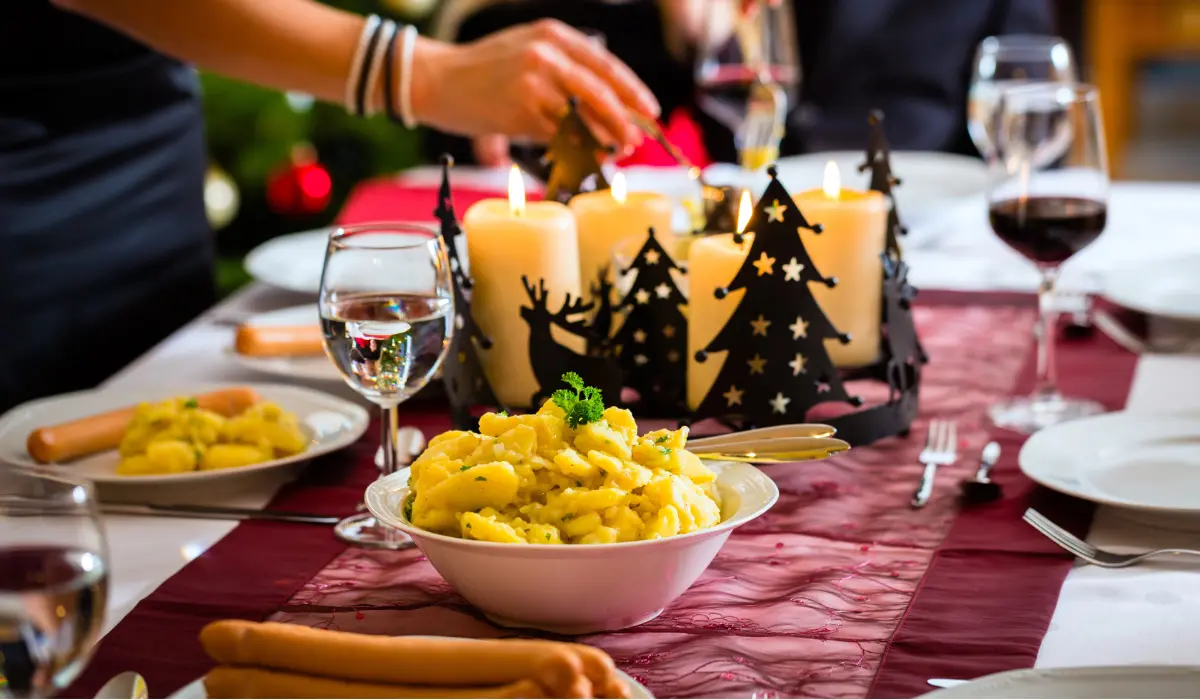 A festive table setting with a bowl of pasta in the foreground, surrounded by decorations, plates, glasses of wine, and lit candles shaped like trees.