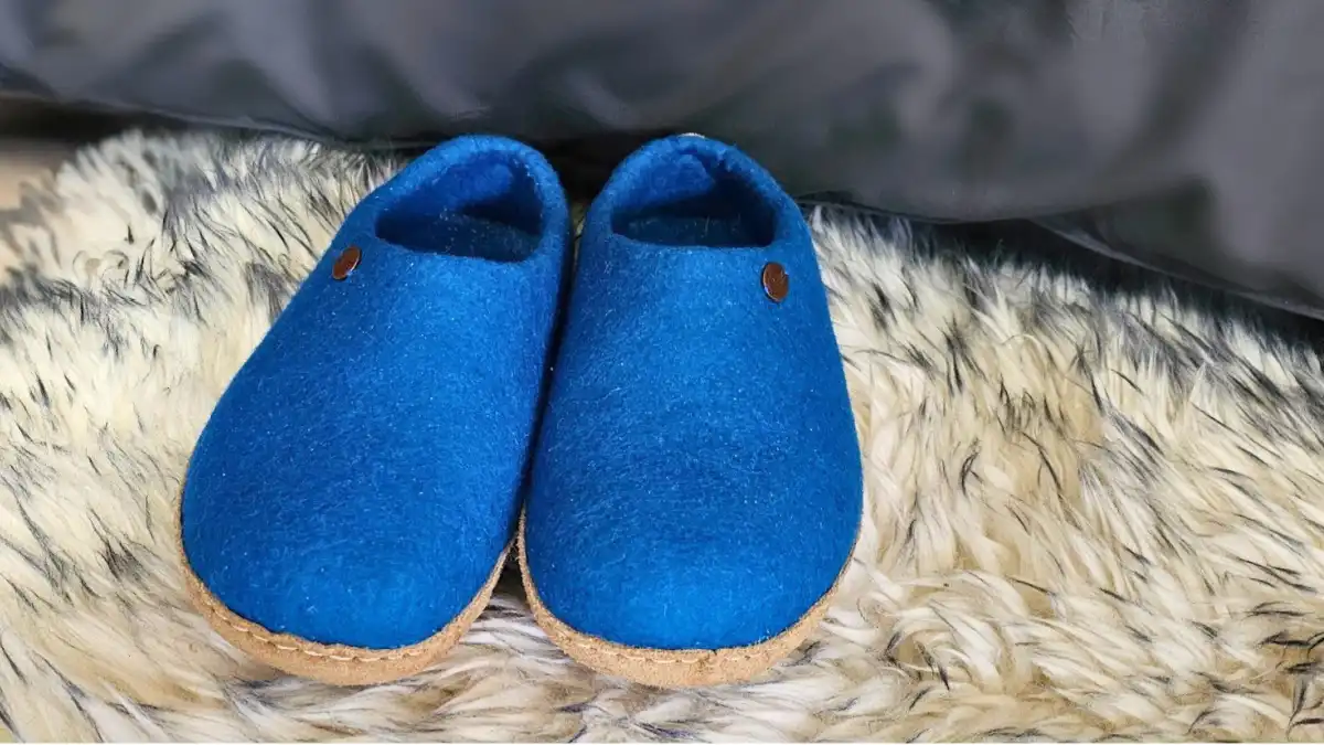Pair of blue WoolFit Footprint slippers, placed neatly on a sheepskin rug beside a bed.
