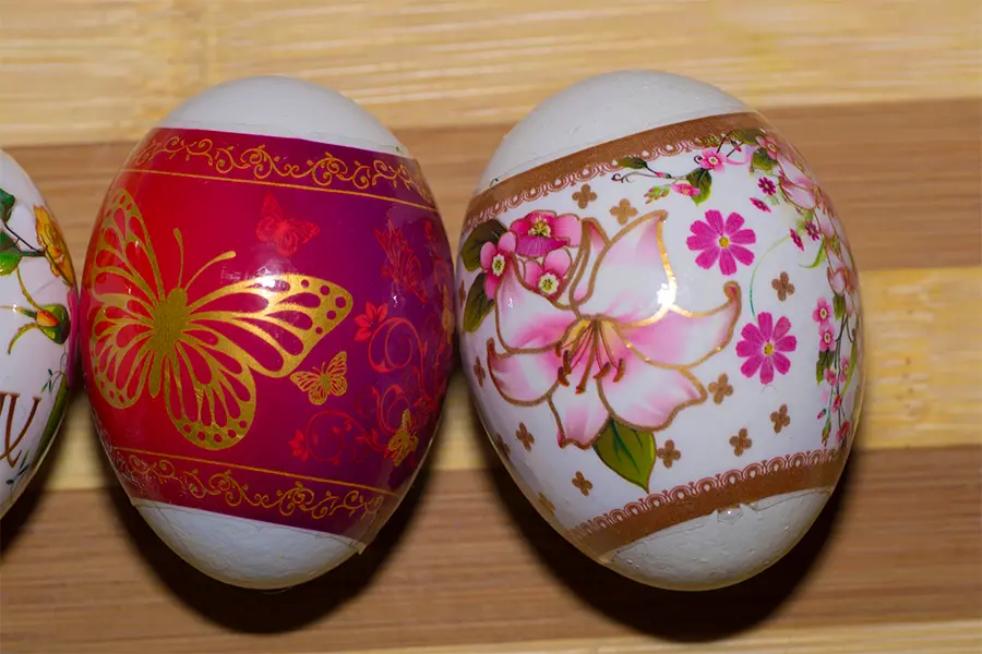 Two decorated eggs; one with a pink and gold butterfly design, the other with pink flowers and green leaves on a white background, placed on a wooden surface.