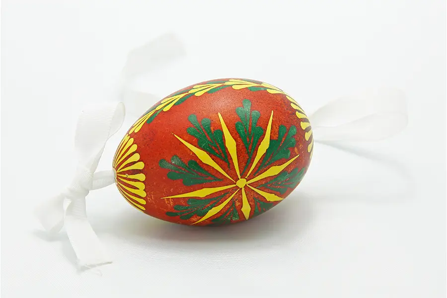 A decorative egg with a red base is adorned with intricate green and yellow patterns. White ribbons are attached to each end, enhancing its presentation.
