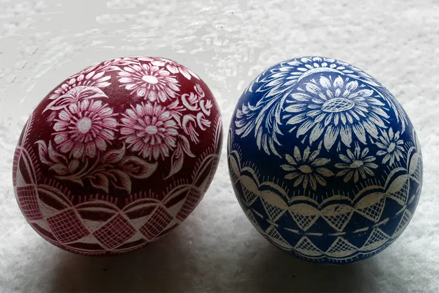 Two intricately decorated eggs with floral patterns; one is maroon, the other is blue, sitting on a textured white surface.