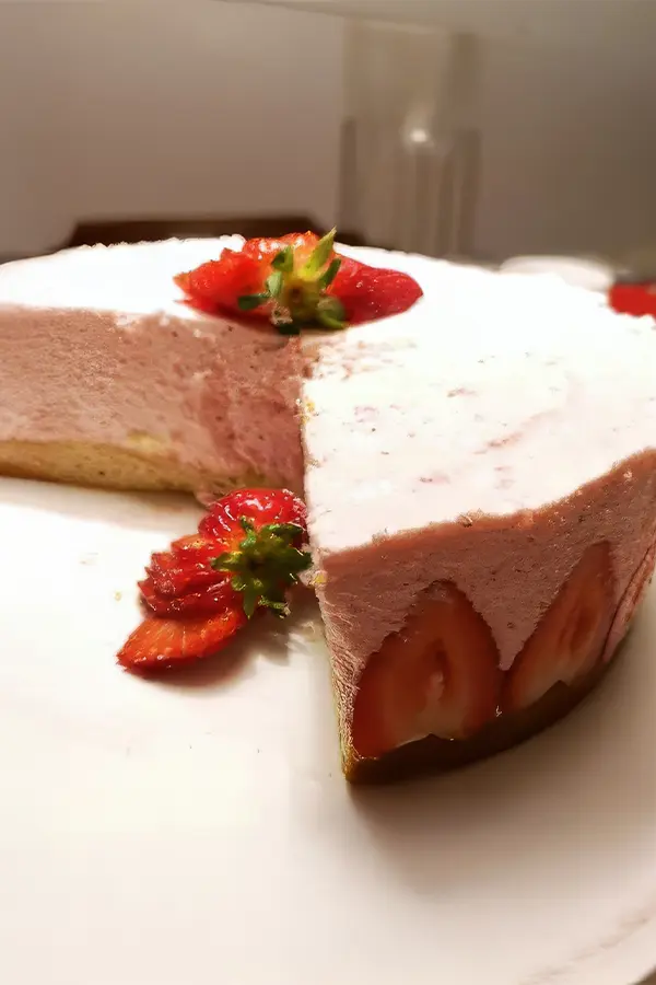 Strawberry mousse cake with a slice removed, revealing strawberries embedded within. Decorated with sliced strawberries on top.