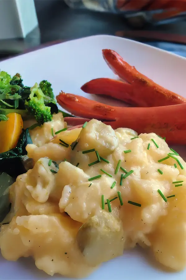A plate with sliced sausage, warm potato salad topped with chives, and a side of mixed steamed vegetables.