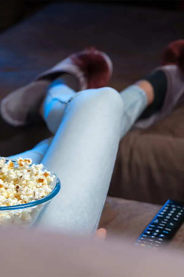 Person in light pants and slippers sits on a couch with a bowl of popcorn and a remote control on the side.