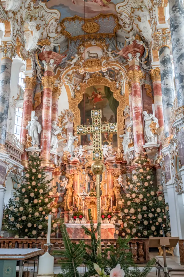 Ornate church altar with a detailed cross, surrounded by Christmas trees and gold accents, featuring a painted ceiling and marble columns.