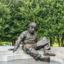 Bronze statue of Albert Einstein seated on a curved bench outdoors, holding a notebook with engraved formulas, surrounded by lush green trees.