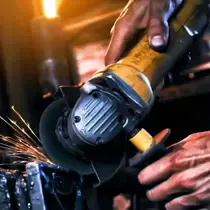 Close-up of a worker's hands using a yellow angle grinder, emitting sparks in a workshop with warm industrial lighting.