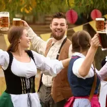 Group of smiling people in traditional Bavarian clothing raising beer mugs in a festive outdoor setting with autumn colors and decorations.