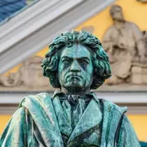 A bronze statue of composer Ludwig van Beethoven with a stern expression, set in front of a yellow historic building in Bonn, Germany.
