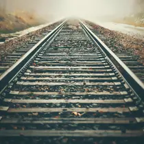 A pair of railroad tracks stretch into the distance, disappearing into a thick, foggy horizon with fallen leaves scattered along the wooden ties.