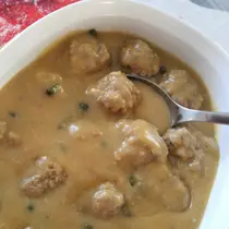 A white bowl filled with German Königsberger Klopse—meatballs in creamy white sauce with capers—being served with a metal spoon.