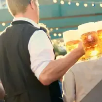 A server dressed in traditional Bavarian attire carrying several large beer steins at an Oktoberfest celebration, with festive lights in the background.