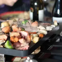 A tabletop raclette grill sizzling with assorted meats, vegetables, and cheese, surrounded by wine bottles and a cozy dining atmosphere.