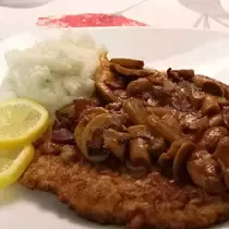A German-style breaded schnitzel topped with mushroom gravy, served with mashed potatoes and lemon slices on a white plate.