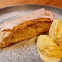 A slice of warm apple strudel filled with spiced apples, served with scoops of vanilla ice cream and dusted with powdered sugar on a grey plate.